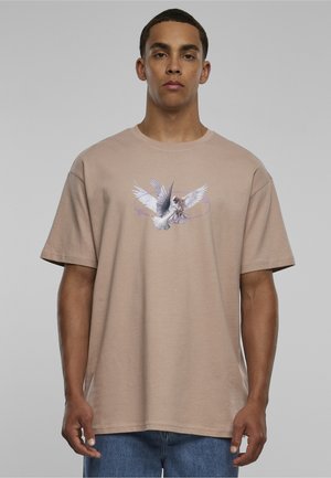 Mężczyzna ubrany w beżowy t-shirt z grafiką dwóch białych gołębi w locie na jednolitym szarym tle.