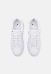 Witte sportieve schoenen met een glad synthetisch bovenwerk, platte veters en een minimalistisch ontwerp. "A/X" logo gedrukt op de tong.
