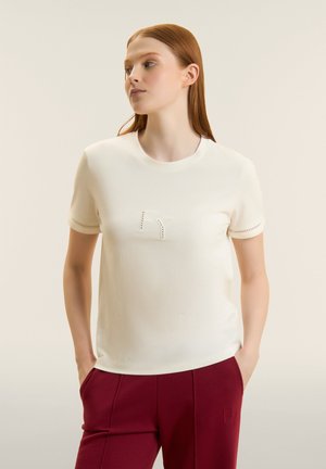 Donna con capelli rossi che indossa una maglietta bianca a maniche corte con dettaglio "FY" e pantaloni bordeaux con le mani nelle tasche, rivolta verso sinistra.