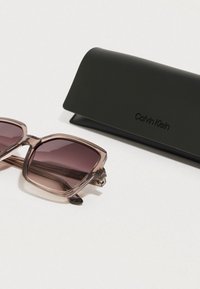 Gafas de sol Calvin Klein con montura transparente color rubor y lentes oscuros degradados, junto con un estuche rígido negro con el nombre de la marca en relieve.