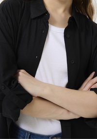 Czarna koszula zapinana na guziki nałożona na białą, prążkowaną bluzkę na ramiączkach, z klasycznym kołnierzykiem, podwiniętymi rękawami i luźnym krojem. Niebieskie dżinsy są częściowo widoczne.