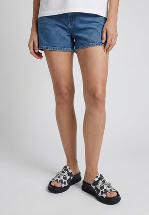 Person trägt blaue Jeansshorts und schwarz-weiße, gemusterte Open-Toe-Slipper mit dicker Sohle und steht vor einem einfarbigen Hintergrund.