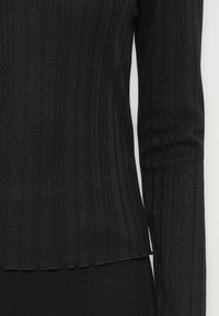 Prim-plan al unui top lung cu mânecă, de culoare neagră, din tricot cu densitate ribbed, evidențiind liniile verticale texturate și un tiv ușor ondulat la talie.