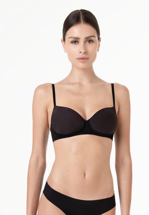Donna con capelli lisci e scuri indossa un reggiseno nero e biancheria intima nera coordinata, in piedi contro uno sfondo bianco semplice.