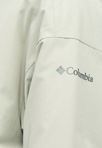 Λεπτομέρεια ανοιχτόγριου μπουφάν της μάρκας Columbia, που δείχνει τις ραμμένες ραφές και το λογότυπο πάνω στο ύφασμα.