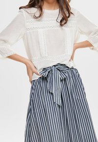 Blouse blanche avec empiècements en dentelle, associée à une jupe rayée marine et blanche avec une ceinture nouée en tissu à la taille et des poches latérales.