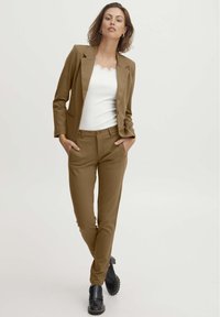 Hnedý blazer cez biely tielko, doplnený o zladené hnedé elegantné nohavice a čierne masívne topánky. Hladká tkanina s priliehavým strihom.