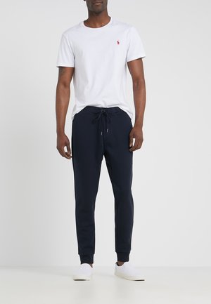 Camiseta de algodón blanca con logotipo rojo en el lado izquierdo del pecho, combinada con pantalones jogger azul marino con cintura ajustable y tobillos elásticos. Zapatillas blancas.