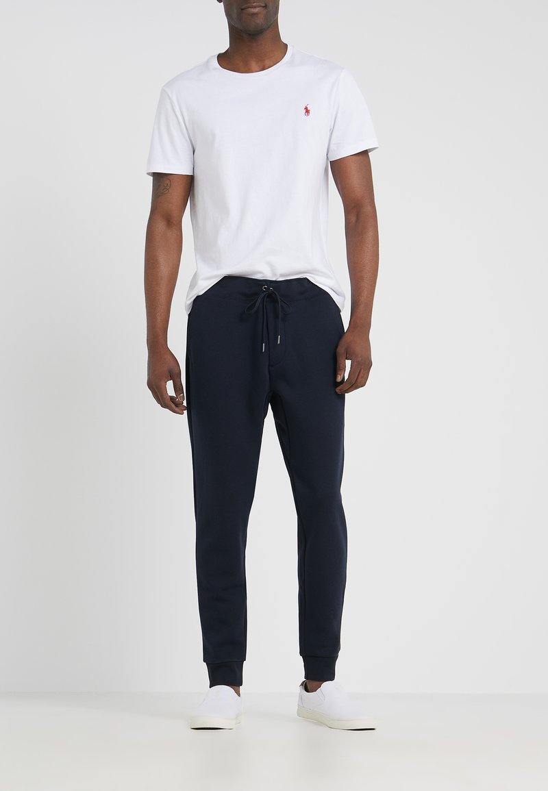 T-shirt en coton blanc avec un logo rouge sur la poitrine gauche, associé à un pantalon de jogging bleu marine avec taille à cordon et chevilles côtelées. Baskets blanches.