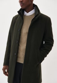 Manteau en laine vert foncé avec un col haut, doté de boutons noirs, de poches latérales et d'une coupe ajustée sur un pull en tricot beige.