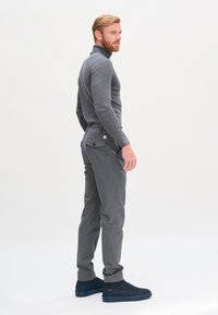 Grauer Rollkragenpullover mit slim fit, kombiniert mit grauen Hosen mit Gesäßtaschen. Das Model trägt dunkle Sneakers. Einfarbiger Hintergrund.
