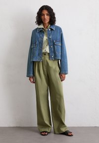 Model stoi przy jasnej ścianie, nosząc krótki dżinsowy żakiet, jasnozieloną koszulę oraz luźne oliwkowe spodnie, do których założyła sandały.