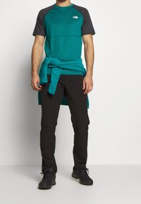 Krótka koszulka sportowa w kolorze zielonkawym i czarnym z okrągłym dekoltem, zestawiona z czarnymi spodniami i ciemnym obuwiem. Koszulka ma logo na piersi.