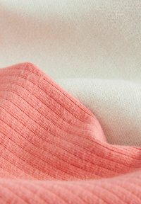 Textílie v koralových a smotanových farbách, s rebrovanou textúrou v koralovej a hladkým povrchom v smotanovej, vrstvené a mierne pokrčené.