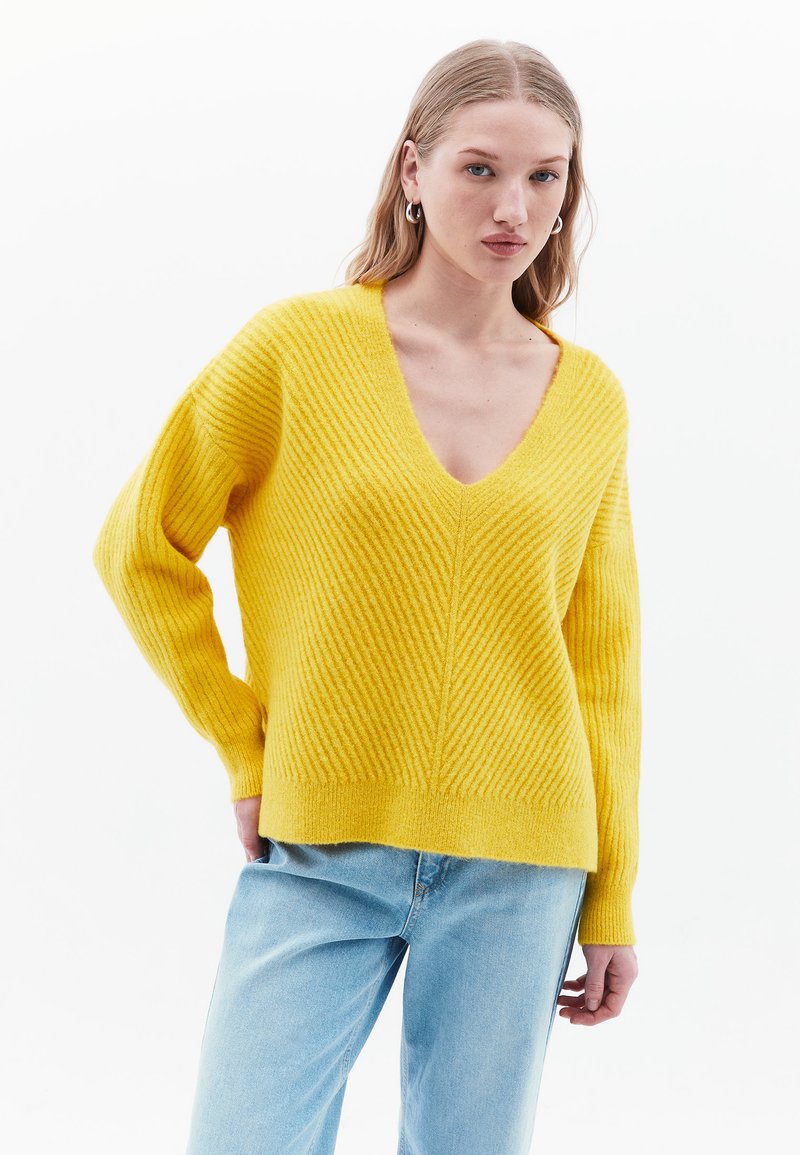 Geel v-hals trui met ribbelstructuur en lange mouwen. Heeft een ontspannen pasvorm en een rechte zoom. Gepaard met lichtblauwe jeans.