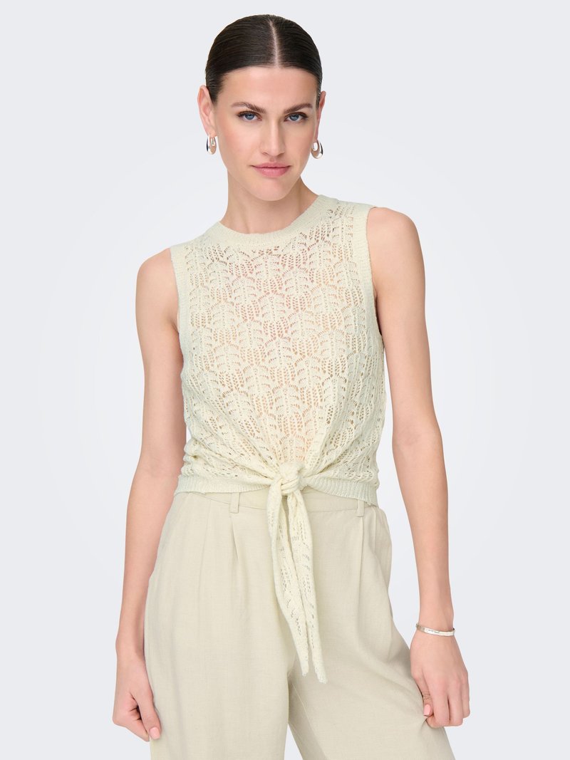 Mujer con top de punto crema sin mangas con lazo delantero y pantalones de talle alto beige claro, de pie frente a un fondo claro y liso.