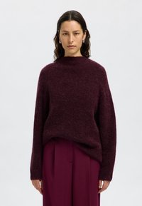 Pull en maille bourgogne avec un col haut, coupe ample et manches longues, assorti à un pantalon plissé. Texture douce, matériau chaud.