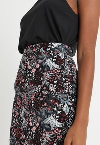 Falda negra con estampado floral, con flores rojas y blancas vibrantes, cinturilla ajustada y tela ligeramente texturizada. Llevada con una blusa negra.