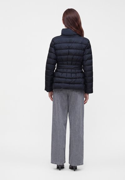 Femme aux longs cheveux bruns, vue de dos, portant une veste matelassée noire, un jean gris à jambes larges et des chaussures noires à talons.