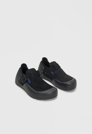 Paire de chaussures noires slip-on avec dessus en daim, semelles en caoutchouc et brides réglables sur le dessus, posées sur une surface gris clair.