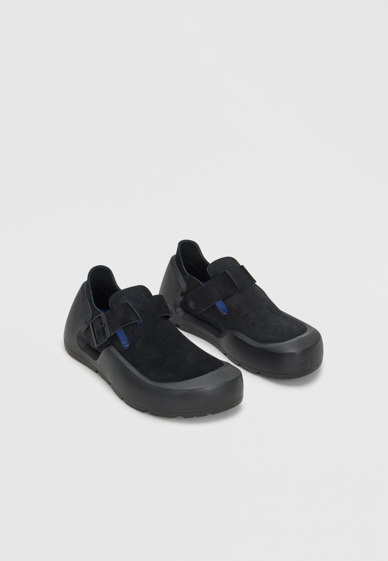Ζευγάρι μαύρα slip-on παπούτσια με σουέντ επάνω μέρος, καουτσούκ σόλες και ρυθμιζόμενα λουράκια στο πάνω μέρος, τοποθετημένα σε ανοιχτό γκρι επιφάνεια.