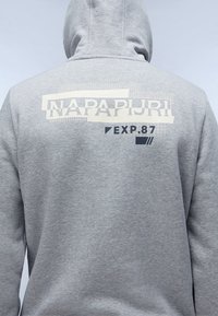 Personne portant un sweat à capuche gris clair avec « NAPAPIJRI » et « EXP. 87 » imprimés en blanc et noir dans le haut du dos.