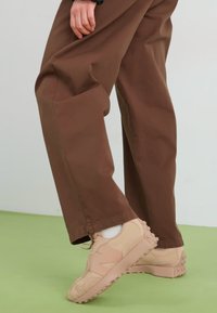 Beige sneakers med teksturert stoff og gummisåler, kombinert med løse brune bukser, står på et grønt gulv mot en hvit bakgrunn.