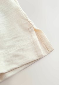 Nahaufnahme eines cremefarbenen Stoffsaums mit verstärkten Nähten und einer kleinen seitlichen Schlitzdetailaufnahme auf hellem Hintergrund.