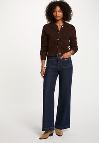 Brauner, taillierter Cardigan mit Rundhalsausschnitt und Knopfverschluss, kombiniert mit dunkelblauen, weiten Jeans und sandfarbenen, hohem Stiefeletten.