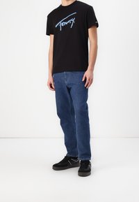 Černé bavlněné tričko s modrou grafikou, kombinované s modrými džínami a černými teniskami. Tenisky mají texturovaný, sportovní design.