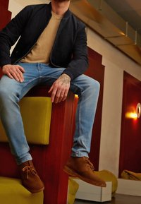 Dark suede bomber jacket over a beige shirt, light blue jeans, and brown lace-up shoes, seated on a wooden structure with yellow accents.