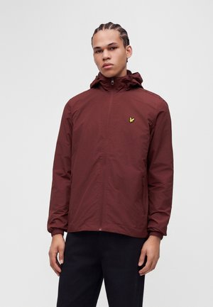 Jeune homme aux cheveux tressés portant une veste à capuche bordeaux avec un logo d'oiseau jaune sur la poitrine et un pantalon noir, debout devant un fond uni.