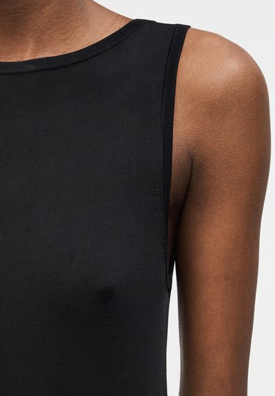 Nahaufnahme einer Personenschulter und des oberen Oberkörpers, die ein ärmelloses schwarzes Oberteil aus strukturiertem Stoff vor einem schlichten Hintergrund trägt.