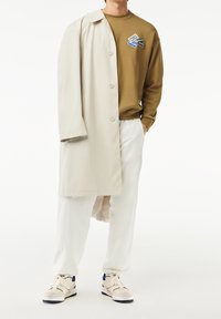 Beige dlouhý kabát, olivový svetr s grafickými akcenty, bílé kalhoty a světlé tenisky. Má knoflíky a uvolněný střih.