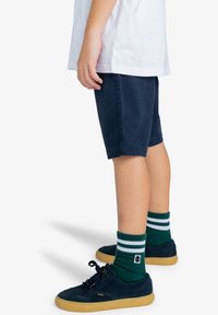Marineblauwe shorts, wit t-shirt, donkergroene gestreepte sokken en marineblauwe schoenen met tanzolen. Zichtbare hand op het shirt, staande positie.