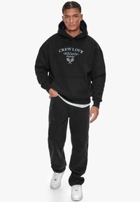 Svart hettegenser med teksten "CREW LOVE athletic", fremre kenguru-lomme og hvite detaljer. Kledd med svarte løse bukser og hvite sneakers.