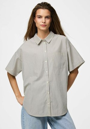 Ung kvinde med langt hår iført en løs, kortærmet, knappet, stribet skjorte og lyseblå jeans, stående med den ene hånd på hoften.