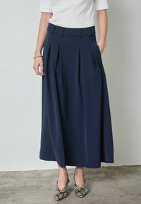 Falda midi plisada azul marino con cinturilla alta y bolsillos laterales, confeccionada en tela suave, combinada con tacones slingback con estampado de serpiente.