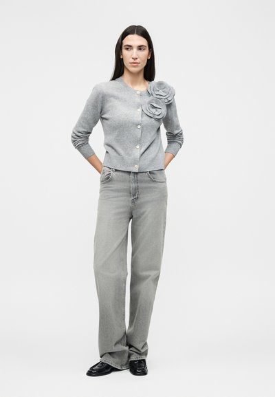 Γυναίκα που στέκεται με τα χέρια πίσω από την πλάτη, φορά γκρι κουμπωτή ζακέτα με δύο υφασμάτινα λουλούδια και άνετο γκρι τζιν με μαύρα παπούτσια.