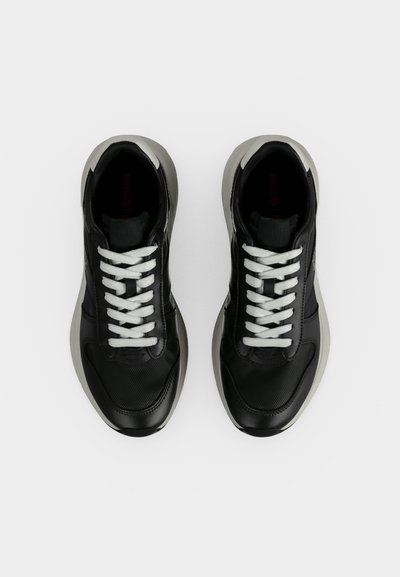 Paire de baskets noires avec des lacets blancs et des semelles blanches épaisses vues de dessus sur un fond blanc uni.