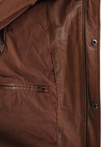 Brun jacka inuti som visar tygfoder, dragkedjeficka och en del av ett knäppt läderöverdrag med synliga sömnadsdetaljer.