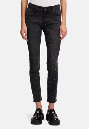 Woman wearing black slim-fit jeans with small silver studs and black chunky loafers, standing against white background.