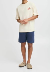 T-shirt créme à coupe décontractée avec un graphic rouge, associée à un short bleu marine et des chaussures slip-on beiges. Matière en coton, design décontracté.