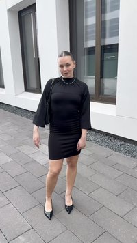 O femeie îmbrăcată într-o rochie neagră și cu tocuri negre stă pe o trotuar pavimentat lângă o clădire albă cu feronerie maro de dimensiuni mari.