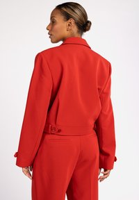 Rode op maat gemaakte blazer met lange mouwen, schoudervullingen en een ceintuur in de taille. Heeft twee knopen bij de manchetten en een gladde afwerking.