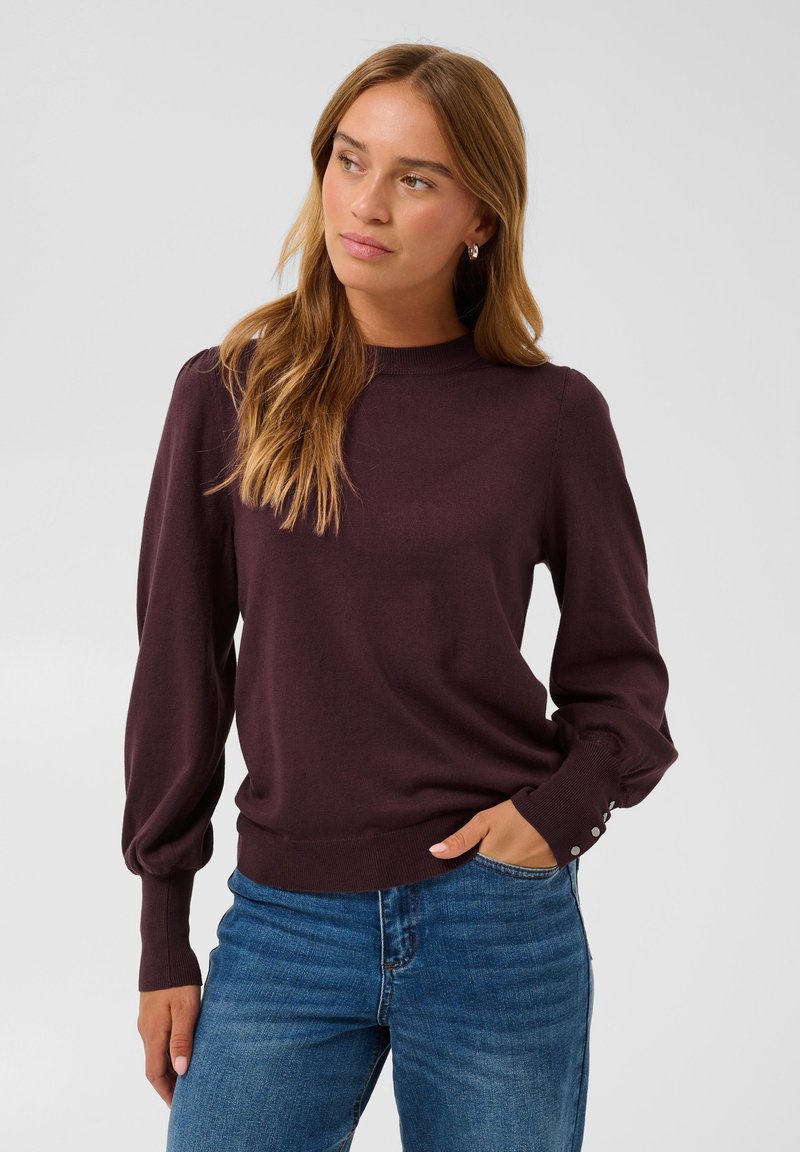 Femme aux longs cheveux châtain clair portant un pull violet foncé à manches longues et un jean bleu, debout avec une main dans la poche, regardant de côté.