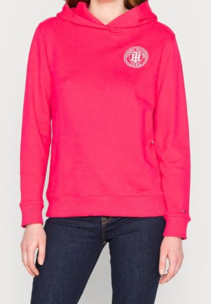 Personne portant un sweat à capuche rose vif avec le logo Tommy Hilfiger sur la poitrine et un jean bleu foncé sur un fond clair uni.