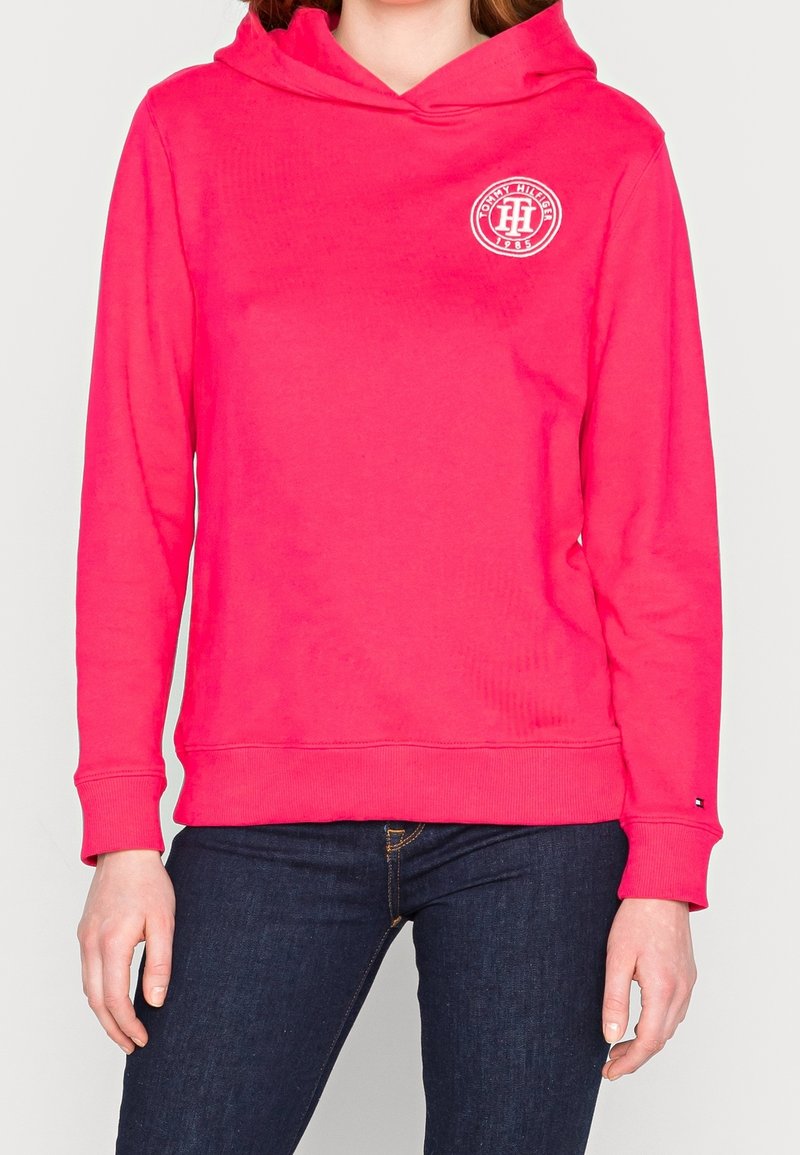 Personne portant un sweat à capuche rose vif avec le logo Tommy Hilfiger sur la poitrine et un jean bleu foncé sur un fond clair uni.