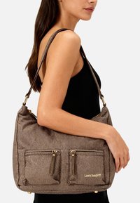 Borsa marrone con texture, dotata di due tasche frontali con cerniera, dettagli in metallo dorato e una tracolla singola. Logo visibile sul davanti.