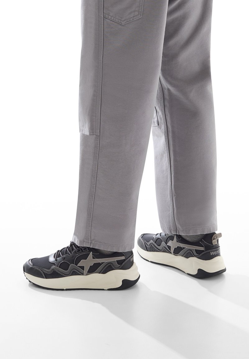 Sneaker sportive nere e grigie con un design ondulato, caratterizzate da un accento a forma di stella, una tomaia testurizzata e una suola bianca spessa, abbinate a pantaloni grigi.